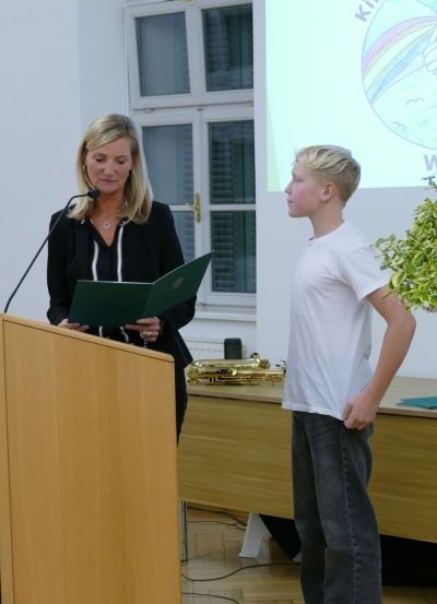 Eine Frau steht an einem Rednerpult mit einem Mikrofon und schaut einen Jungen an, der neben ihr steht. Der Junge schaut in Richtung Publikum. Hinter ihnen ist eine Projektionsleinwand mit einer Präsentation.