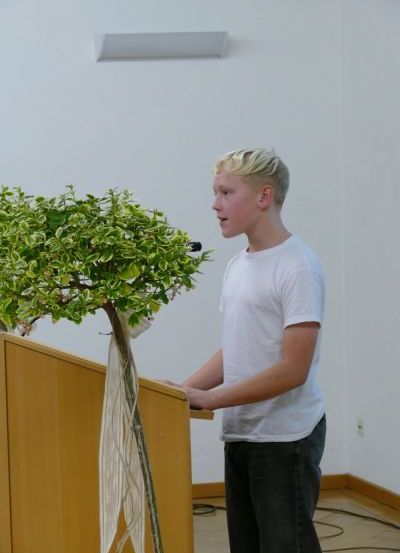 Ein junger Junge steht an einem Podium und spricht in ein Mikrofon, mit einer dekorativen Pflanze dahinter.