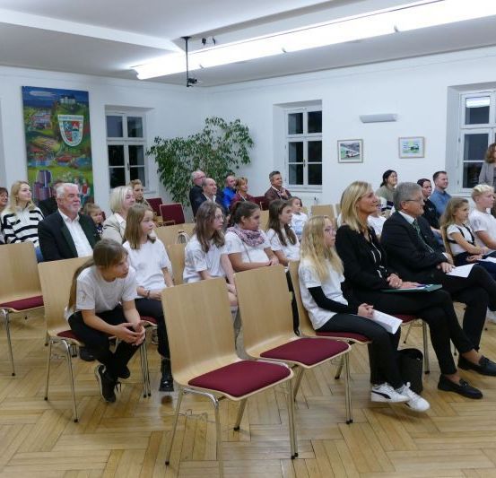 Eine Gruppe von Menschen, darunter Erwachsene und Kinder, sitzen in Stühlen in einem Raum. Sie hören wahrscheinlich einer Präsentation zu.