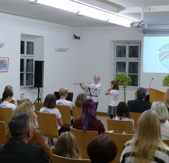 Ein Klassenzimmer mit einer Projektionsleinwand, die das Logo einer Organisation zeigt. Zwei Kinder spielen Musikinstrumente, während eine Gruppe von Menschen zusieht.