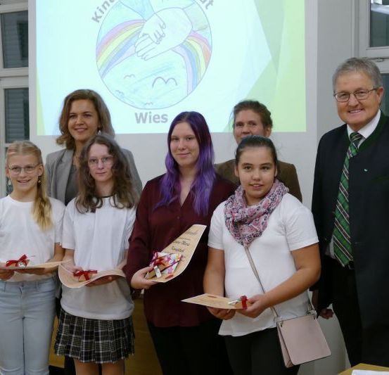 Eine Gruppe von Menschen posiert für ein Foto. Sie halten Urkunden und lächeln. Dahinter befindet sich eine Projektionsleinwand mit einer Zeichnung eines Regenbogens und Hände.