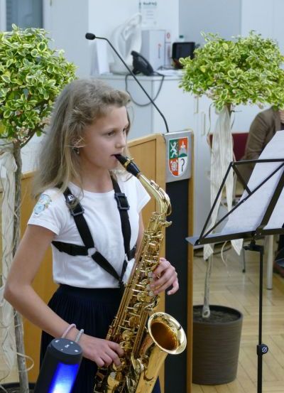 Ein junges Mädchen spielt die Saxophon, trägt ein weißes Shirt und schwarze Träger, mit zwei Topfpflanzen und einem Notenständer im Hintergrund.