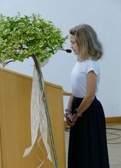 Ein junges Mädchen in einem weißen T-Shirt und einer schwarzen Schürze steht neben einem Podium mit einem Mikrofon und hält eine mit Bändern verzierte Pflanze. Der Ort scheint ein formeller Anlass zu sein.