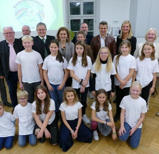 Eine Gruppe von Menschen, darunter Erwachsene und Kinder, posiert für ein Foto. Die Kinder tragen weiße T-Shirts. Einige Erwachsene stehen, andere knien. Dahinter befindet sich eine Projektionsleinwand.