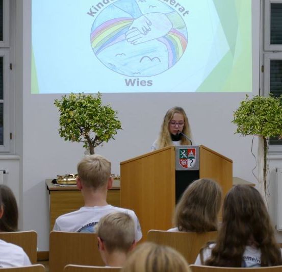 Ein junges Mädchen hält eine Rede an einem Rednerpult in einem Klassenzimmer mit einer Projektionsleinwand hinter ihr. Die Leinwand zeigt ein farbenfrohes Bild und Text. Mehrere Schüler sitzen auf Stühlen und hören aufmerksam zu.
