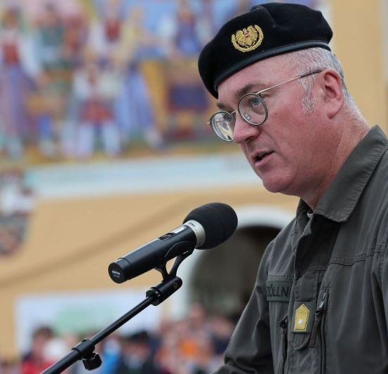 Ein Mann in Uniform spricht in ein Mikrofon im Freien, mit einer Menschenmenge im Hintergrund.