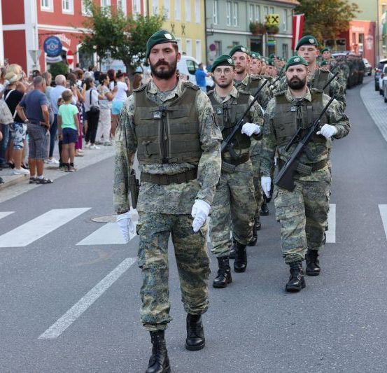 Eine Reihe von Soldaten in Tarnuniformen marschiert eine Straße hinunter, wobei Zuschauer vom Bürgersteig zusehen.