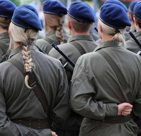 Eine Gruppe von Militärangehörigen steht in Formation, trägt grüne Uniformen und blaue Barette und hält Gewehre.