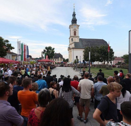 Eine Menschenmenge versammelt sich für eine Veranstaltung auf einem Stadtplatz mit einer Kirche, Flaggen und einem Militärfahrzeug. Die Menschen tragen verschiedene Kleidungsstücke, und einige halten Schilder.