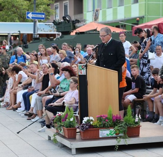 Ein Mann spricht an einem Podium mit Blumen, während ein Militärfahrzeug hinter einer Menschenmenge geparkt ist. Uniformierte Personen sind anwesend.