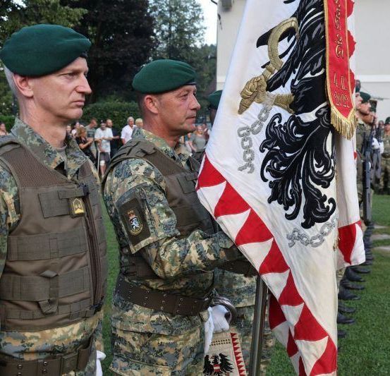 Zwei Soldaten in Uniform halten eine Flagge in einem Außenbereich. Andere Soldaten stehen in Formation hinter ihnen. Die Soldaten tragen Tarnuniformen.
