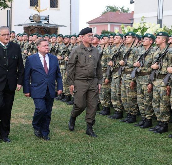Mehrere Männer in Anzügen und einer Militäruniform gehen zwischen einer Reihe von Soldaten mit Gewehren auf einem Feld.