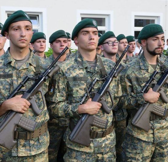 Eine Reihe von Soldaten in Tarnuniformen hält Gewehre und steht vor einem Gebäude mit weißen Fenstern.
