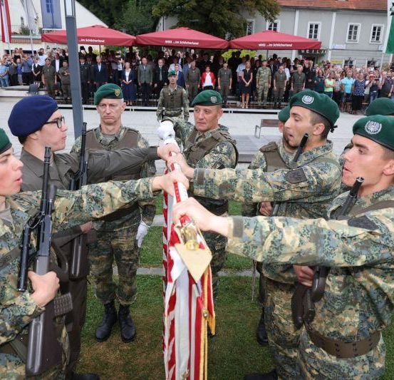 Militärangehörige in Uniform halten eine Flagge während einer Zeremonie, während ein Publikum hinter roten Ständen beobachtet.
