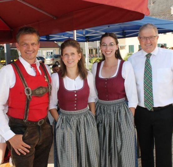 Drei Menschen in traditioneller Kleidung stehen unter einem Regenschirm. Ein Mann in roten Hosenträger und eine Frau in einem weißen Oberteil stehen neben einer Frau in einem roten Oberteil und Rock. Dahinter steht ein blaues Zelt.
