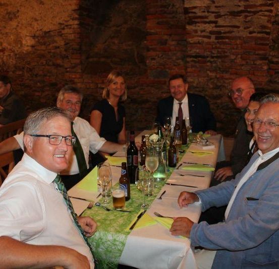Eine Gruppe von Menschen sitzt an einem langen Tisch und lächelt für ein Foto. Sie tragen Brillen, Krawatten und Anzüge. Flaschen, Weingläser und eine Blumenvase stehen auf dem Tisch.