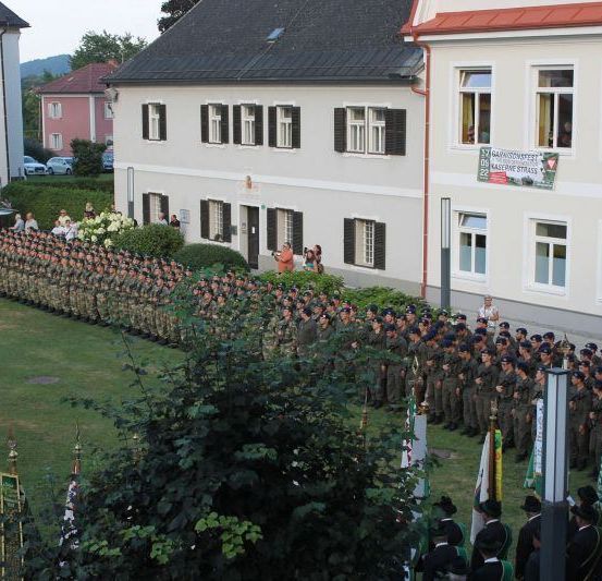 Eine Militärparade auf einem Rasenfeld mit Gebäuden und Bannern im Hintergrund.
