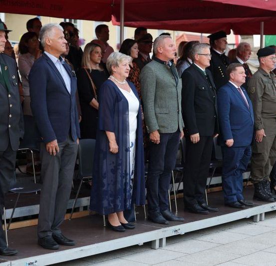 Menschen in formeller Kleidung stehen in einer Reihe unter roten Schirmen, wahrscheinlich bei einer Zeremonie.