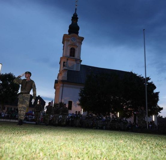Ein Soldat salutiert unter einem bewölkten Himmel vor einer Kirche, vor der sich viele Menschen versammelt haben.