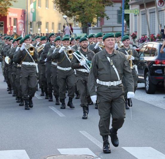 Eine Blaskapelle mit Trompeten und Posaunen führt eine Parade durch die Straße. Zuschauer und Fotografen halten das Ereignis fest.