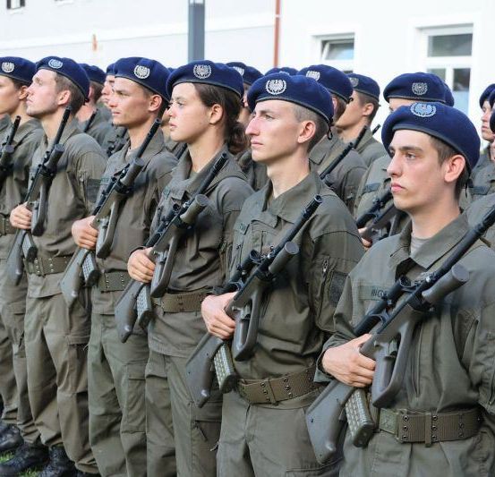 Eine Reihe von Militärangehörigen in grünen Uniformen, die Gewehre halten, auf Gras stehend, mit Gebäuden im Hintergrund.