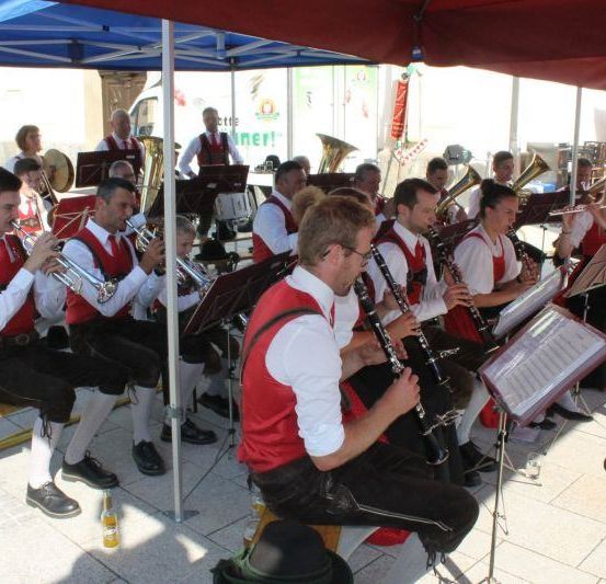 Eine Gruppe von Musikern in traditioneller Kleidung spielt unter einem Zelt verschiedene Instrumente wie Trompeten, Posaunen und Klarinetten.
