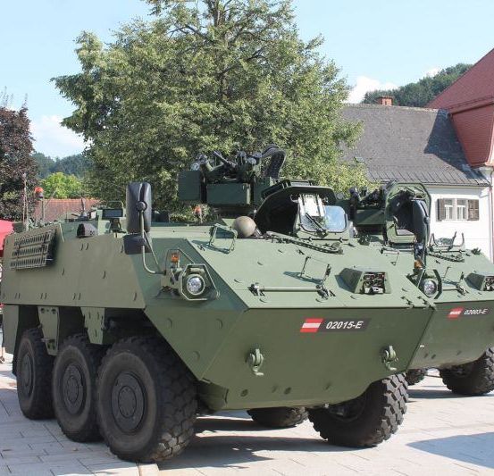 Zwei Militärfahrzeuge sind auf einem gepflasterten Bereich geparkt. Ein Fahrzeug hat die Nummer 02015-E. Dahinter befinden sich Gebäude, Bäume und Menschen.