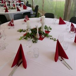 Ein runder Tisch mit weißer Tischdecke, eingerichtet für eine Dinnerparty mit roten Servietten, Kerzen und Gläsern. Zwei weitere Tische sind teilweise im Hintergrund zu sehen.
