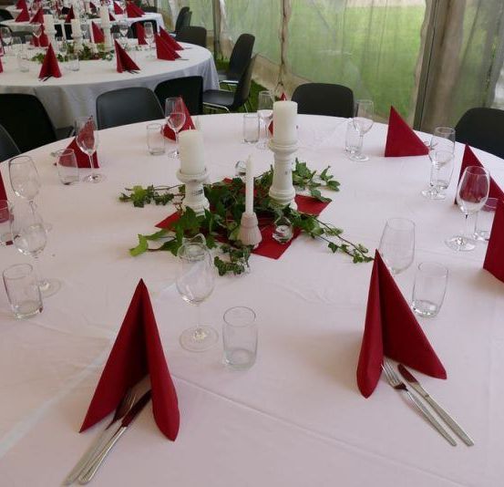 Ein runder Tisch mit weißer Tischdecke, eingerichtet für eine Dinnerparty mit roten Servietten, Kerzen und Gläsern. Zwei weitere Tische sind teilweise im Hintergrund zu sehen.