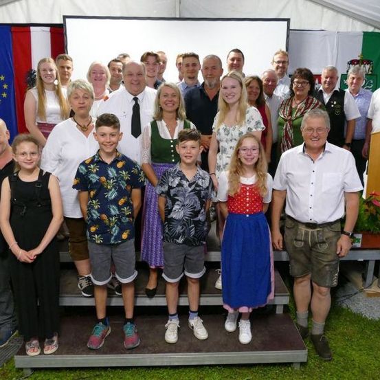 Eine Gruppe von Menschen steht auf einer Bühne in einem Zelt mit Fahnen dahinter. Einige tragen traditionelle deutsche Kleidung. Ein Podium steht rechts und eine weiße Leinwand dahinter.