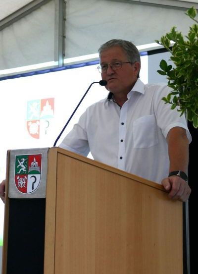 Ein Mann steht an einem Rednerpult mit einem Mikrofon. Er trägt ein weißes Hemd und eine Brille. Hinter ihm befindet sich ein Bildschirm mit einem Logo.