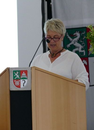 Eine Frau mit Brille steht an einem Rednerpult mit einem Mikrofon und hält eine Rede. Hinter ihr ist eine Wand mit einer Fahne und einem weißen Banner.