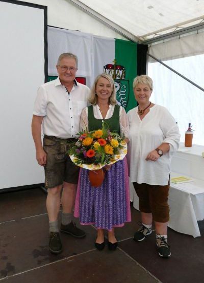 Drei Menschen stehen in einem Zelt und lächeln. Eine Frau hält einen Blumenstrauß. Hinter ihnen befindet sich ein weißes Banner mit einer grünen Flagge.