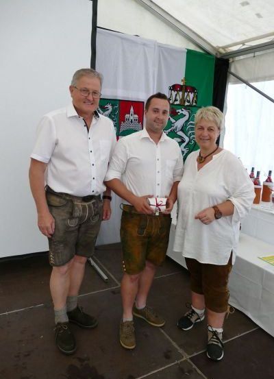 Drei Menschen stehen in einem Zelt, lächeln und einer hält ein Geschenk. Sie tragen traditionelle deutsche Kleidung. Hinter ihnen ist ein weißer Hintergrund mit einer Flagge.