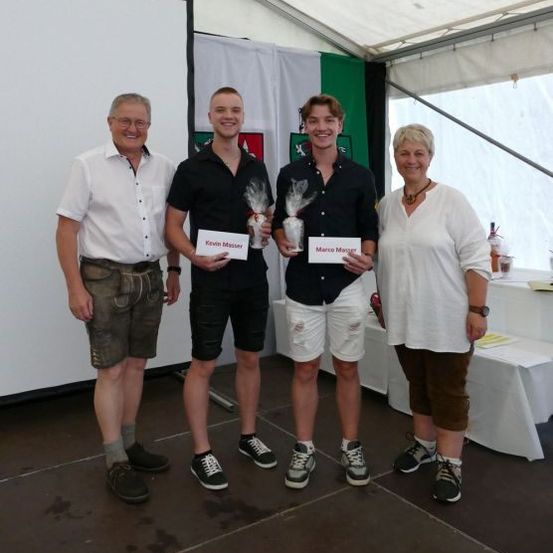 Vier Personen stehen in einem Zelt, zwei Männer in der Mitte halten Auszeichnungen, während eine Frau rechts in die Kamera lächelt. Ein Banner hängt hinter ihnen.