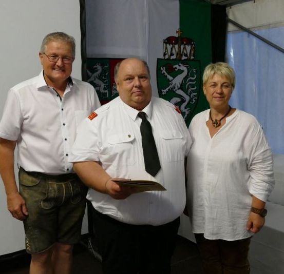 Drei Personen stehen in einem Raum mit einer weißen Tischdecke auf einem Tisch und Emblemen an der Wand. Ein Mann auf der linken Seite trägt eine Brille und grüne Shorts. Der Mann in der Mitte hält ein Stück Papier. Die Frau auf der rechten Seite trägt eine Halskette.