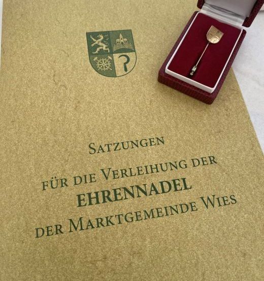 Ein Dokument mit dem Titel 'Satzungen' liegt auf einem Tisch offen. Eine rote Schachtel mit einer goldenen Nadel daneben. Die Nadel trägt ein Logo.
