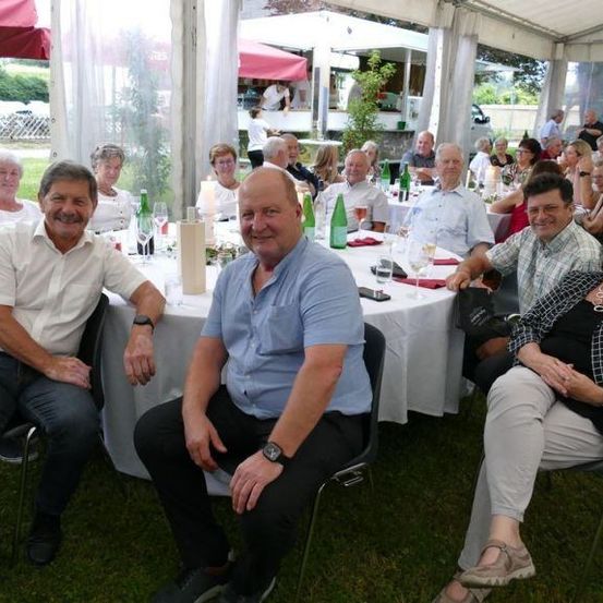Eine Gruppe von Menschen sitzt um einen Tisch in einem Zelt mit grünen Flaschen und Weingläsern. Sie tragen alle Freizeitkleidung.