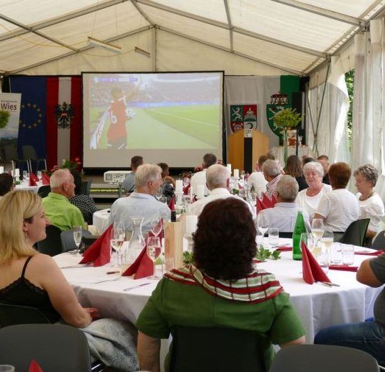 Mehrere Menschen sind für eine Veranstaltung unter einem Zelt versammelt. Eine Beamerleinwand zeigt ein Fußballspiel, und die Teilnehmer sind an Tischen Platz genommen.