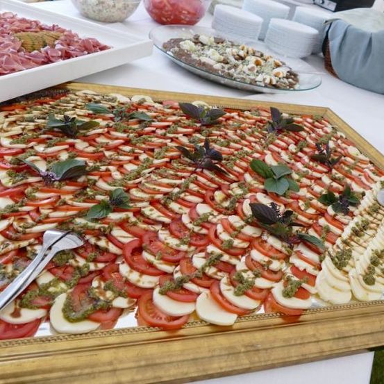 Ein großer Tisch ist mit mehreren Essensplatten bedeckt, darunter ein Herzstück aus geschnittenen Tomaten und Käse mit Pesto. Verschiedene andere Gerichte und Fleischsorten sind ebenfalls auf dem Tisch.