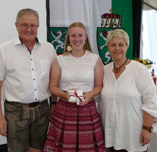 Drei Personen stehen zusammen vor einer grün-weißen Flagge und lächeln für ein Foto. Eine Frau hält ein Geschenk, und eine andere Frau trägt eine Halskette. Hinter ihnen stehen Flaschen auf einem Tisch.