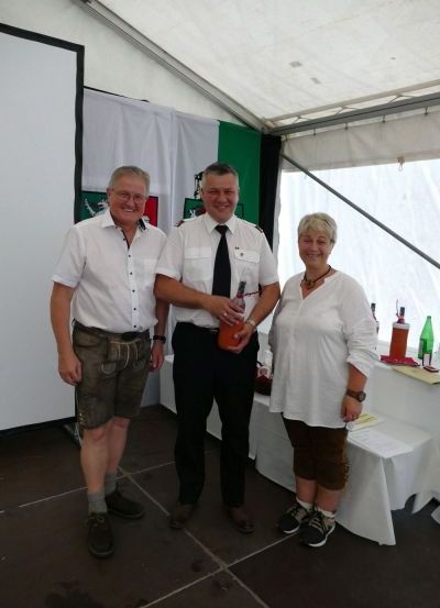 Drei Personen stehen in einem Zelt. Zwei Männer tragen weiße Hemden, einer mit Krawatte und einer in Shorts. Die Frau trägt ein weißes Oberteil. Sie alle lächeln. Ein Tisch mit Flaschen steht hinter ihnen.