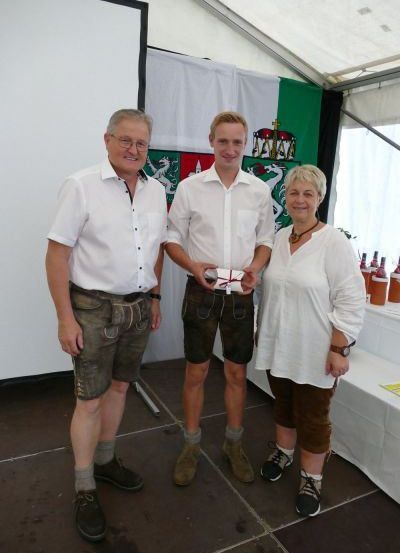Drei Personen stehen eng beieinander in einem Zelt und lächeln. Ein Mann links trägt ein weißes Hemd und Shorts, während der Mann in der Mitte ein Geschenk hält. Eine Frau rechts trägt eine Halskette. Hinter ihnen steht ein Tisch mit Flaschen.
