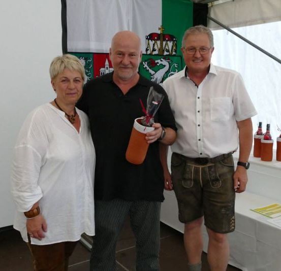 Drei Personen stehen vor einem Tisch mit Flaschen und einer Fahne. Der Mann auf der linken Seite hält ein Geschenk.