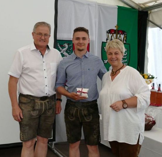 Drei Personen stehen eng beieinander und lächeln für ein Foto. Der Mann auf der linken Seite trägt ein weißes Hemd und grüne Shorts. Der Mann in der Mitte hält ein kleines Geschenk. Die Frau auf der rechten Seite trägt eine weiße Bluse. Hinter ihnen steht ein Tisch mit Flaschen und einer Blumenarrangement.