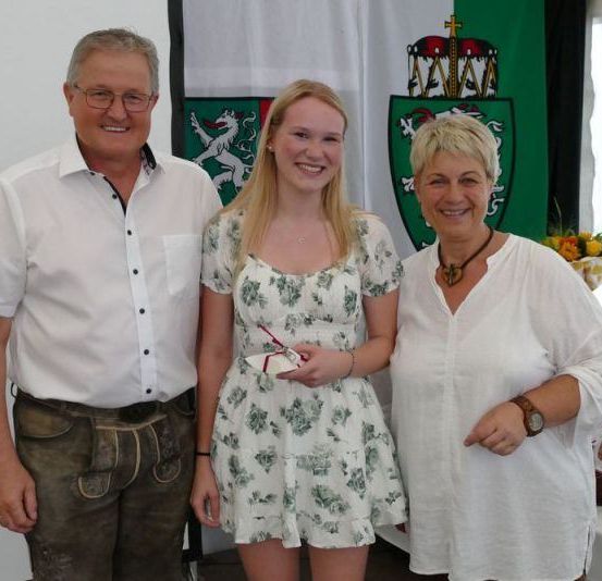 Drei Menschen stehen zusammen und lächeln für ein Foto. Der Mann auf der linken Seite trägt ein weißes Hemd und braune Hosen. Die Frau in der Mitte hält ein kleines Objekt, möglicherweise einen Preis. Die Frau auf der rechten Seite trägt ein weißes Oberteil und eine Halskette. Hinter ihnen steht ein weißer Tisch mit Blumen und Flaschen.
