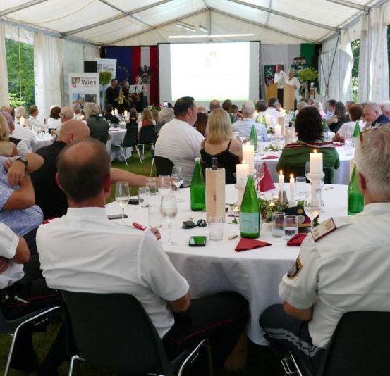 Eine große Gruppe von Menschen sitzt in einem Zelt mit weißen Vorhängen, einer Projektionsleinwand und einer Bühne mit einem Rednerpult und einem Mikrofon. Sie sind in weißen Uniformen gekleidet. Die Tische sind mit weißem Tuch bedeckt und haben Gläser, Flaschen und rote Servietten. Die Menschen schauen auf die Projektionsleinwand.