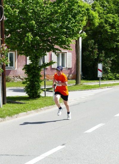 Ein Mann läuft auf einer Straße mit einem Baum und einem Haus im Hintergrund. Er trägt ein oranges Shirt, schwarze Shorts und weiße Schuhe.