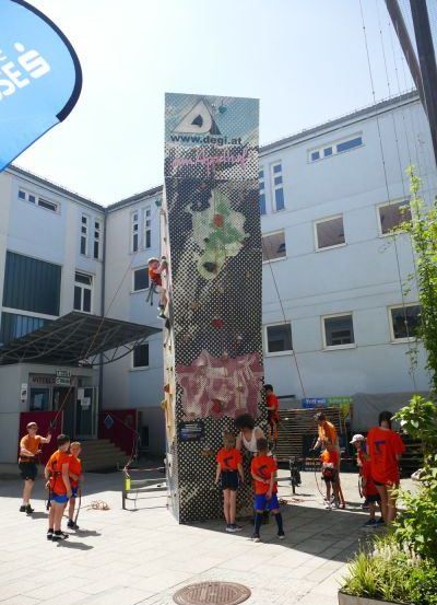 Freiluft-Kletterwand an einem Schulgebäude mit Schülern in orangefarbenen Hemden und Shorts, einige klettern und andere beobachten.