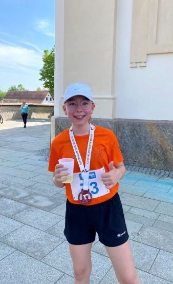 Ein junger Mensch mit einer Medaille um den Hals steht draußen an einem sonnigen Ort, lächelt und hält einen Becher.